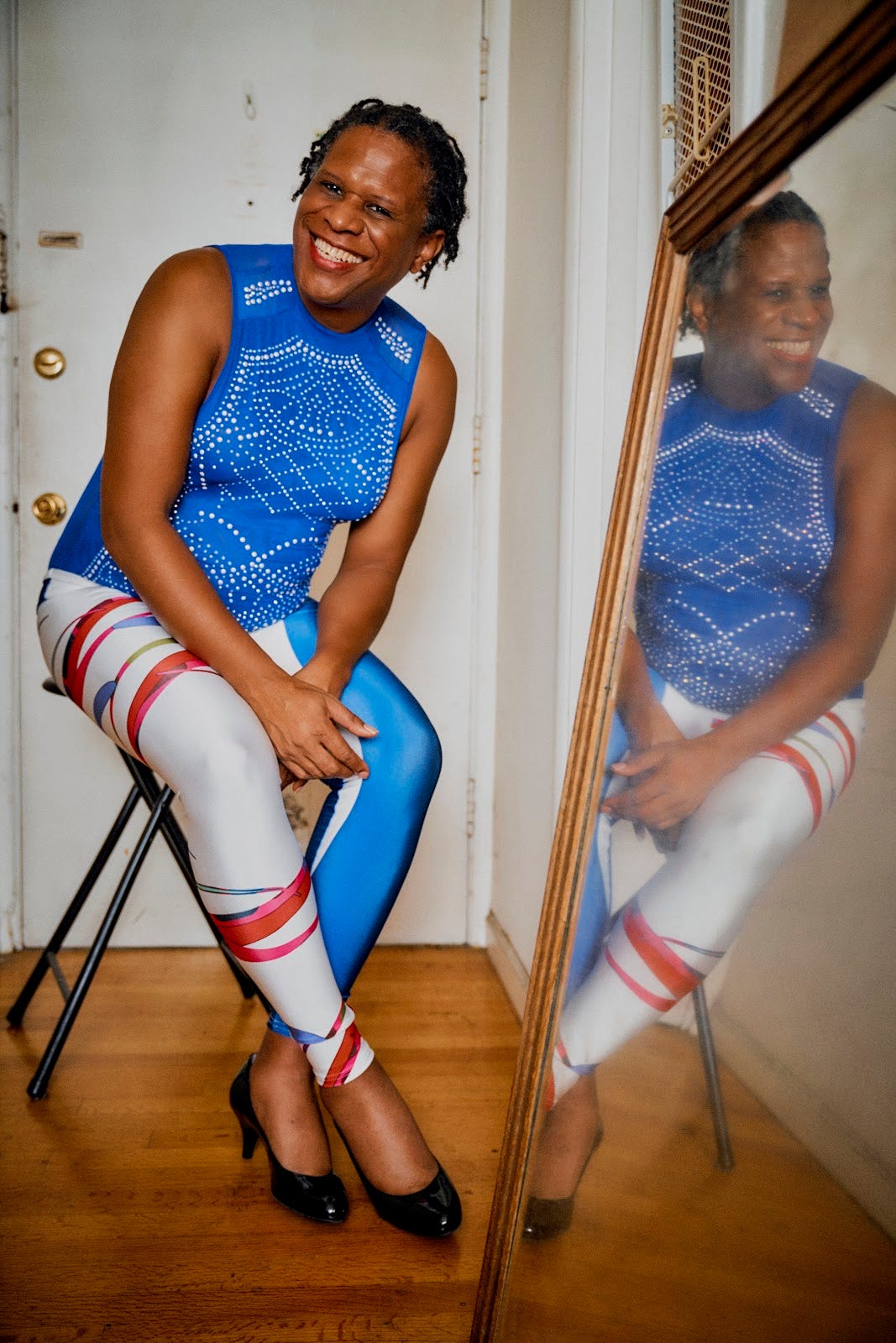 A Black woman with short twists sitting next to a large mirror sitting on the floor and propped up against a wall.