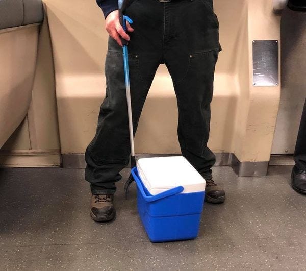 A person from the waist down on a BART train, a white picker-upper in their hand and a blue-and-white cooler on the ground.
