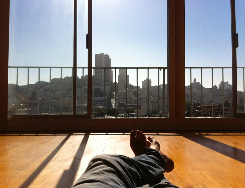 Point-of-view photo of the writer’s legs as he sits in front of a sliding glass window with a view of SF buildings.