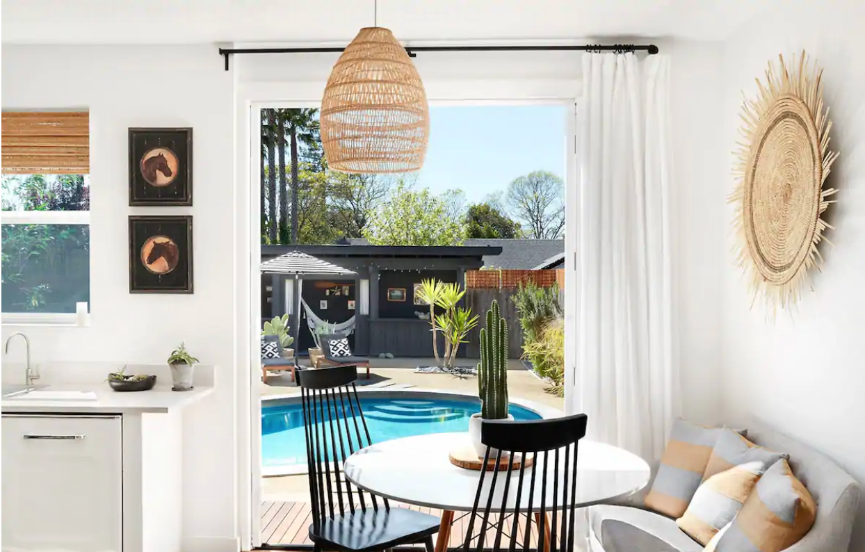 View of a circular pool and a small, gray wooden house, taken through a large door inside a stylish room.