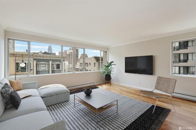 A room with an L-shaped sofa and a wall-mounted flat screen. The window shows buildings from across the street.