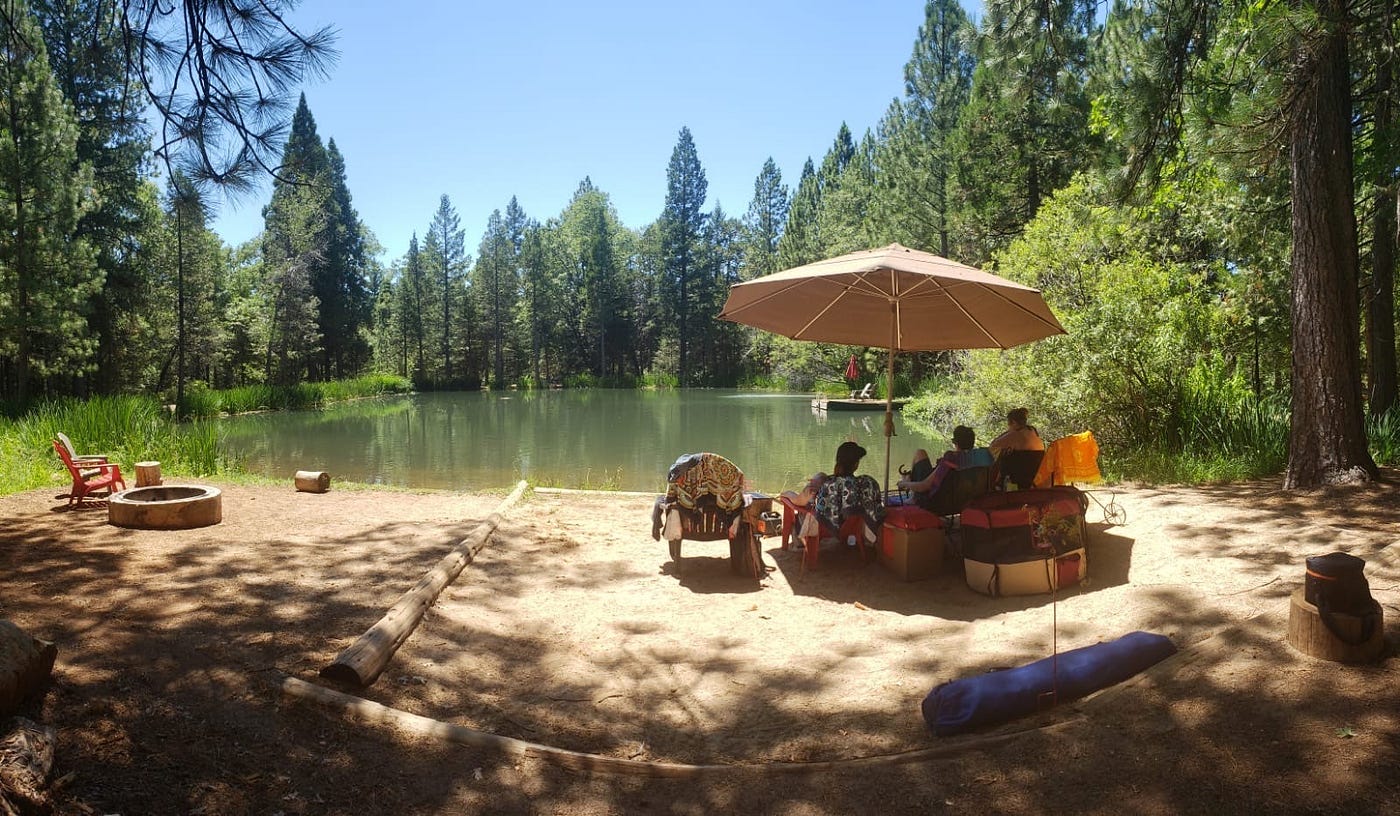 People sitting under a large umbrella on a sandy beach next to a lake.