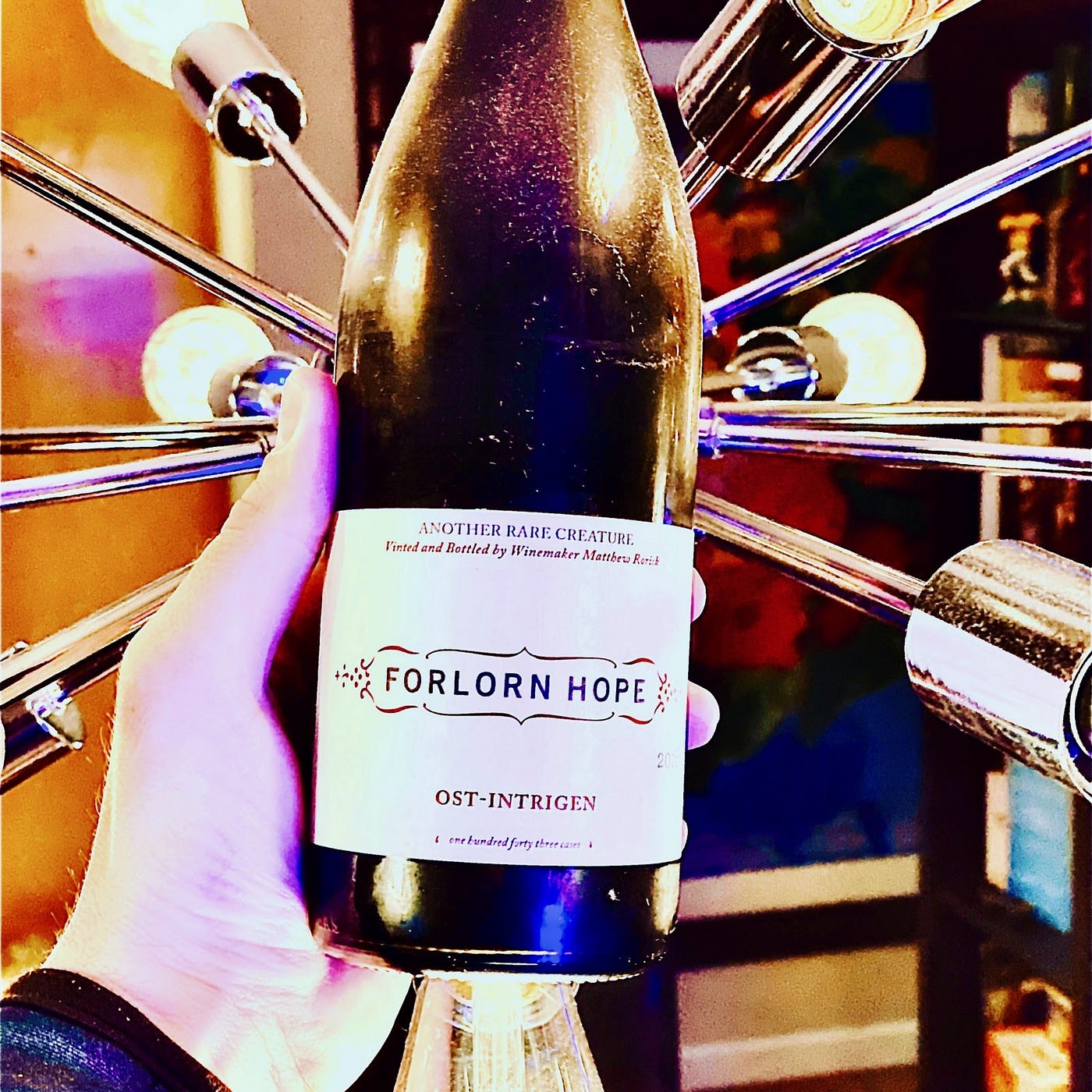 A hand holding a bottle of Forlorn Hope’s Ont-intrigen wine in front of a metallic geometric light fixture.