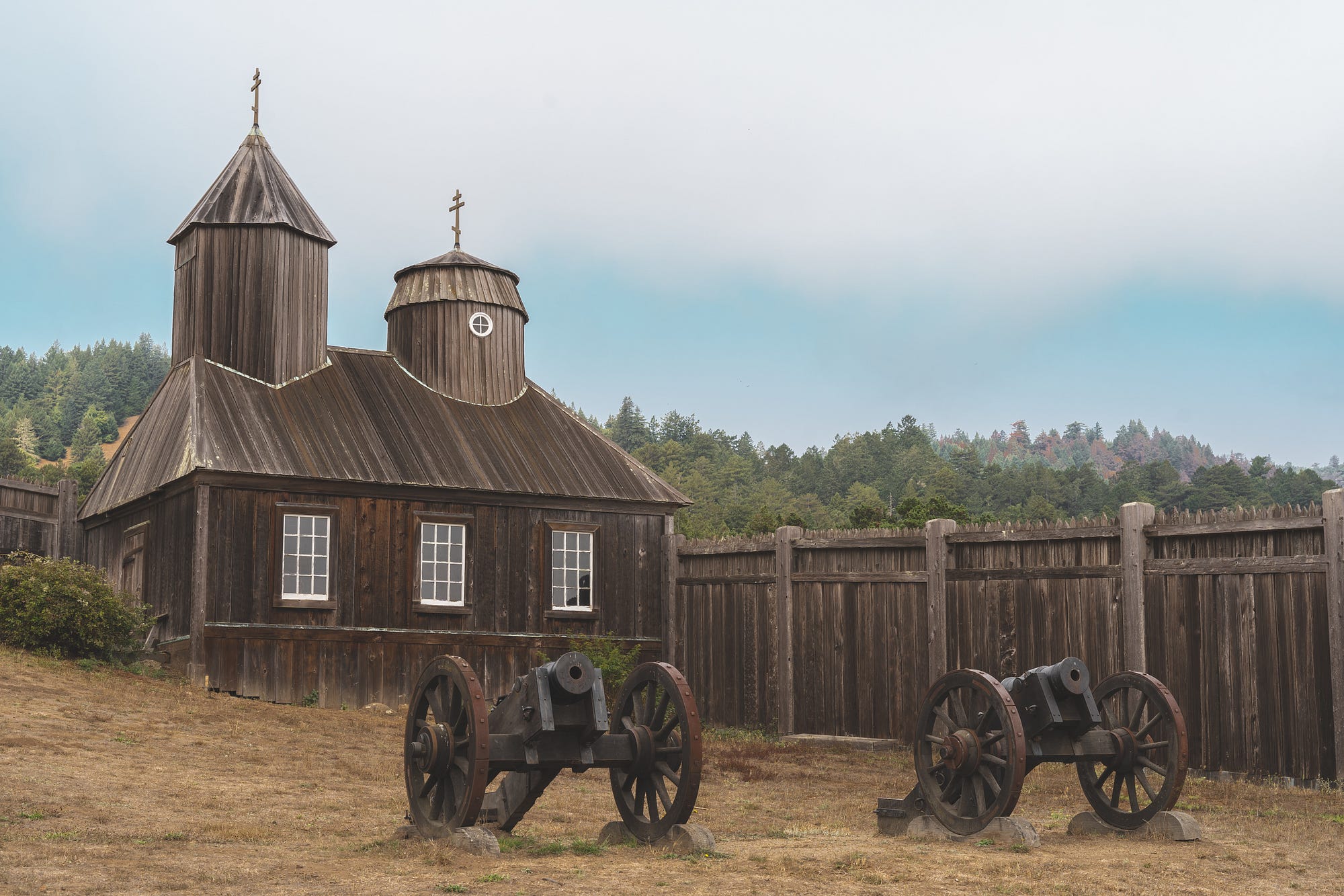 A wooden Russian Orthodox church with two antique cannons in the foreground and a wooden fence in the background.