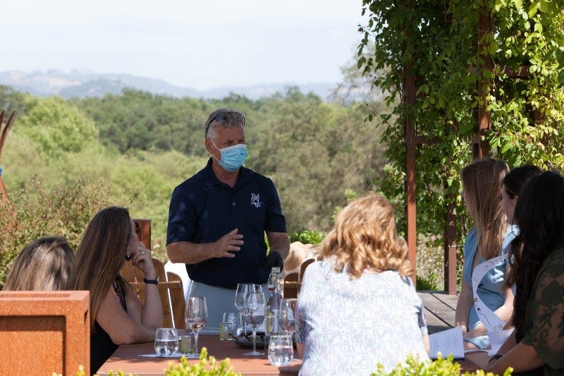 A masked waiter talks to a group of people sitting at a table outside, wine glasses in front of them.