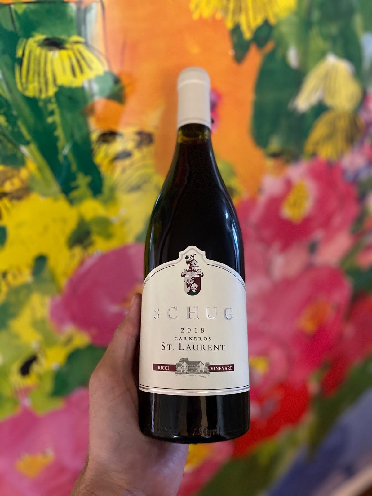 A hand holding up a bottle of Shug’s 2018 Carneros St. Laurent wine in front of floral wallpaper or a painting.