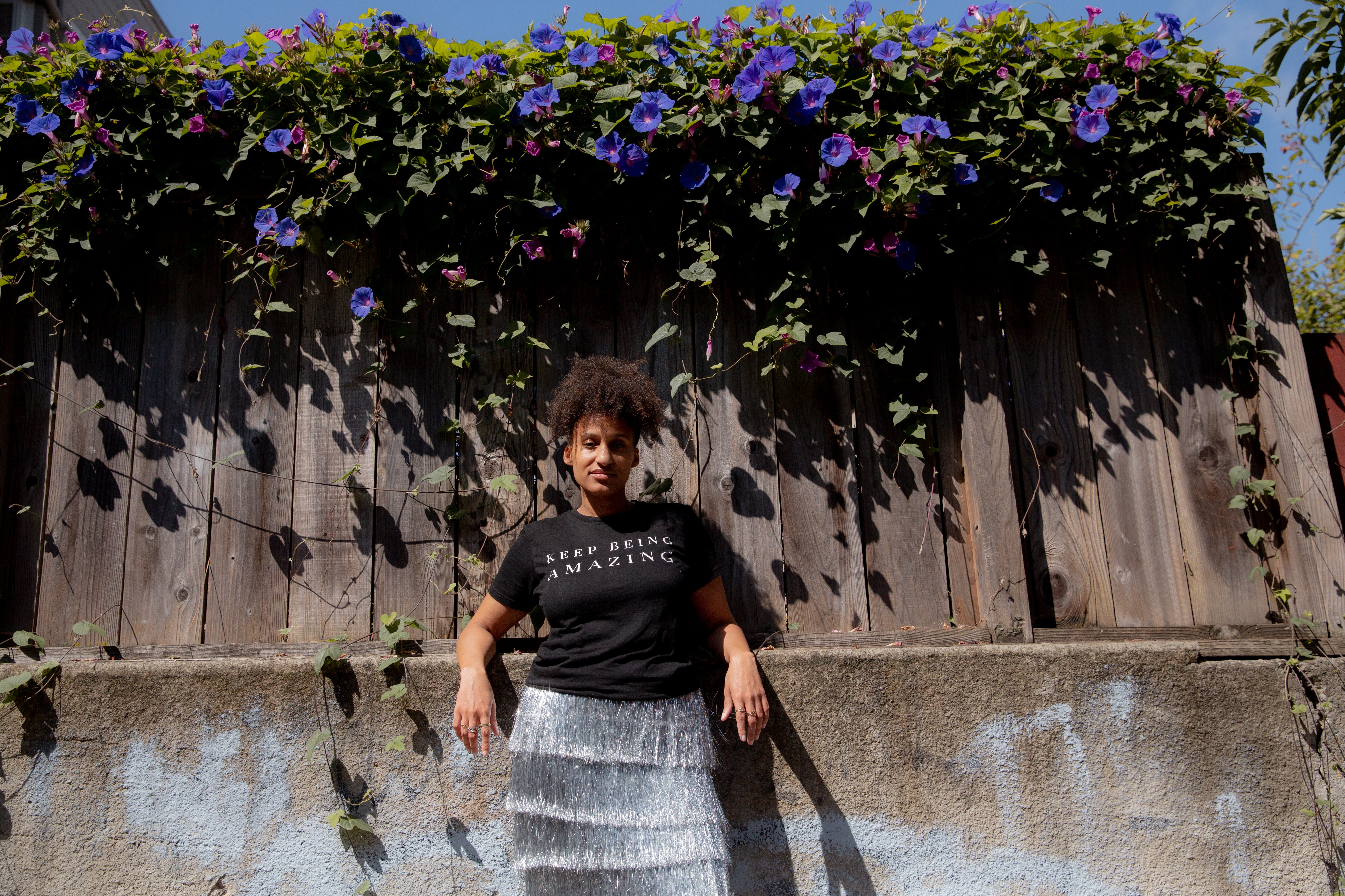 Akilah Cadet leaning backwards on her elbows against a concrete retaining wall. A fence with flowering ivy is above her.