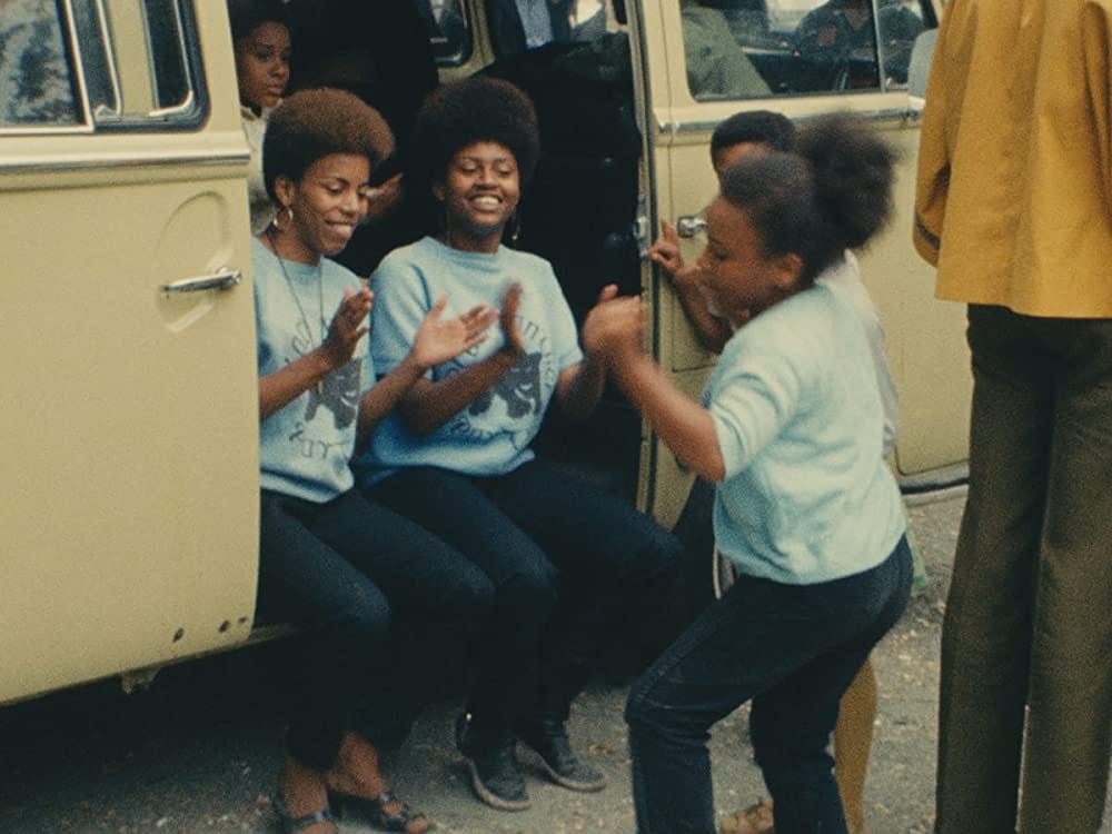Black women sitting on the floor of a van with its doors open, clapping as children dance in front of them.