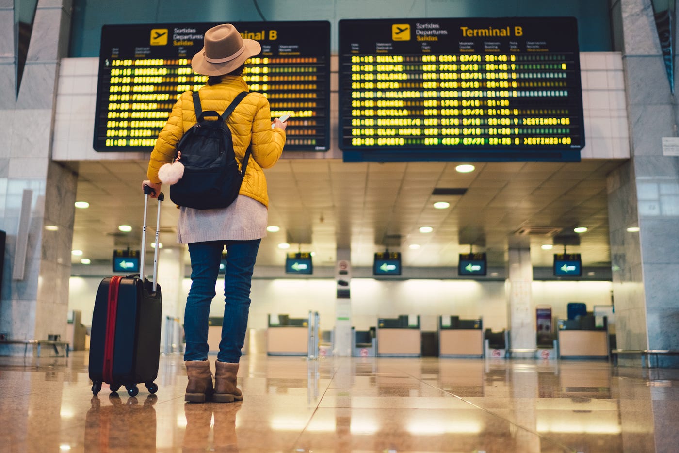 A traveler with a backpack and a rolling suitcase looks at arrivals boards at the airport.
