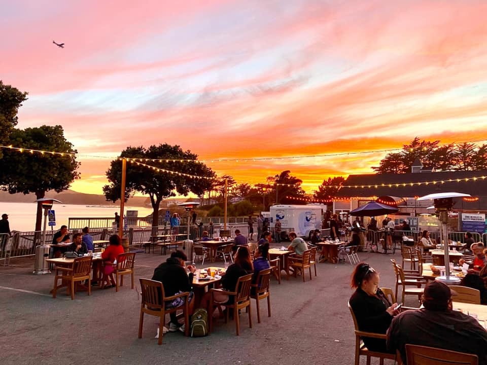Diners sitting at wide-spaced tables under string lights outdoors; the twilight sky has pink, orange, and yellow clouds.