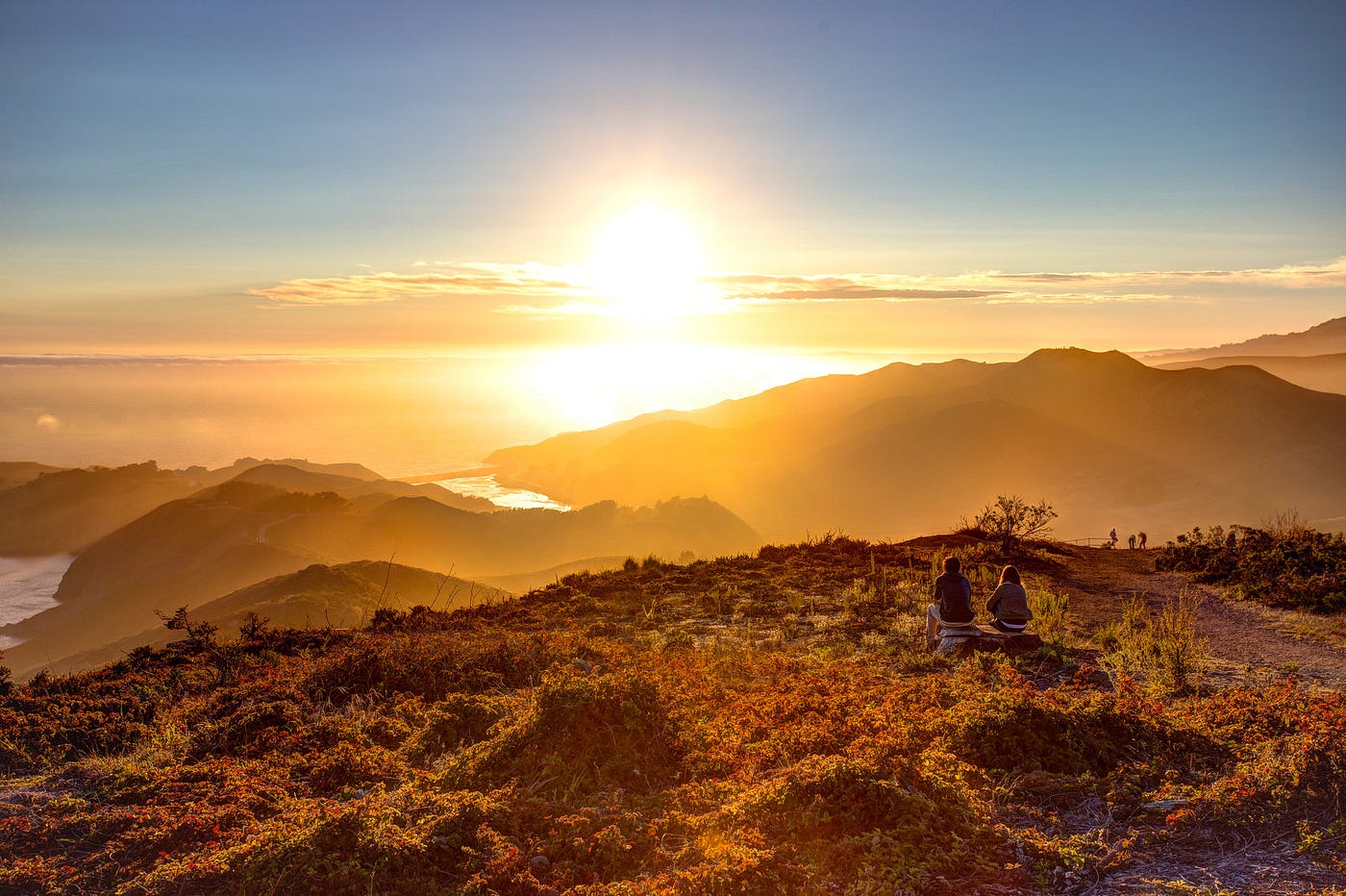 A shot from far away of two people sitting on rocks, watching the sun set over some hills.