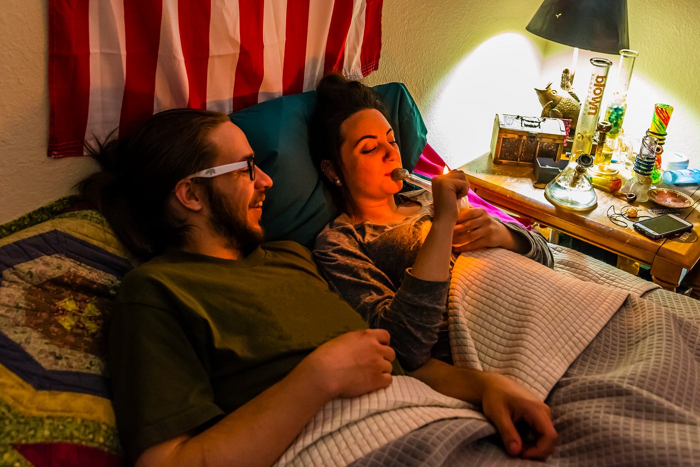 Two young people smoking out of a bong, lying on a bed with a US flag hung on the wall behind them.