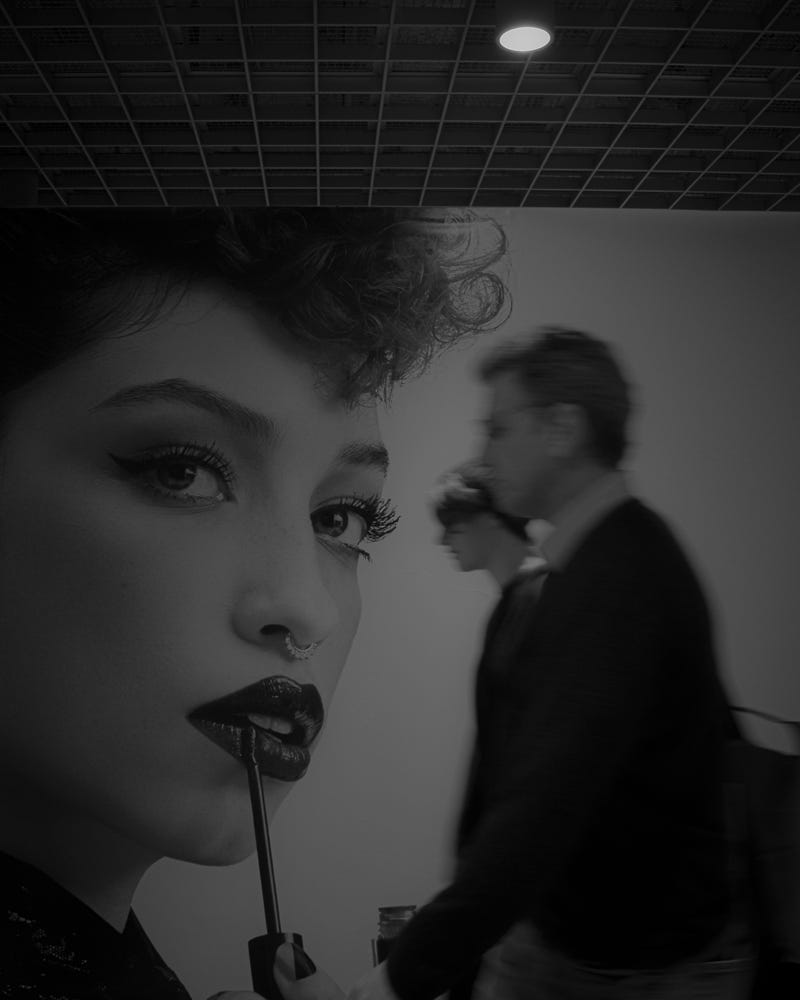 Black-and-white photo of a model’s face with people walking in the background.