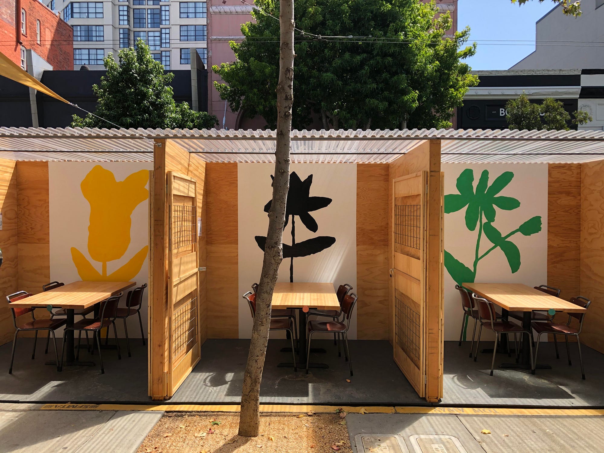 An outdoor dining space with 3 cubicles with dinner tables and flower murals on the wall.