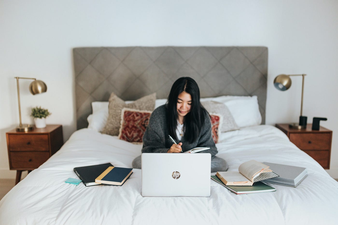 A person with a blanket draped around them sitting in bed working on a laptop, surrounded by books.