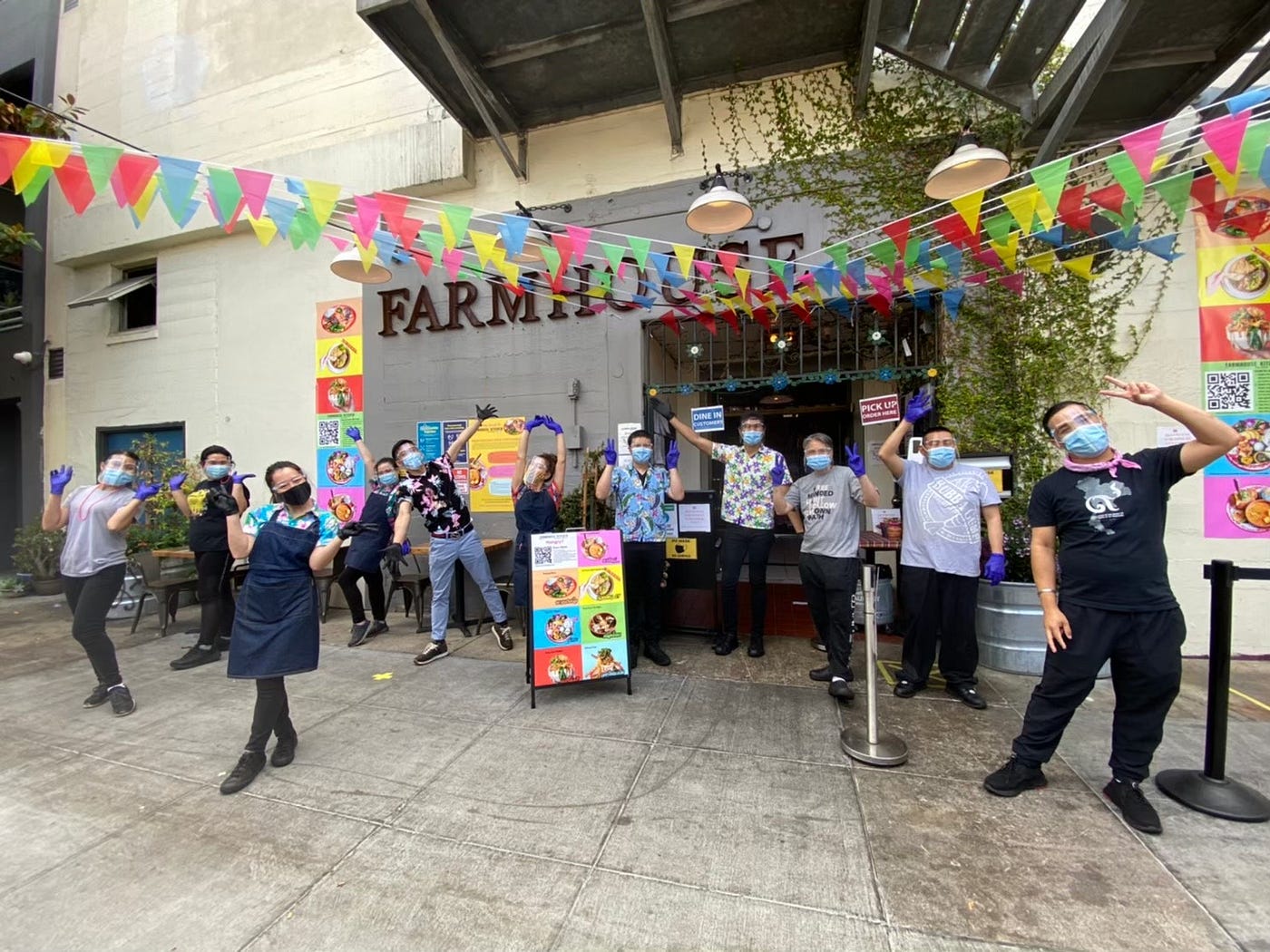 Masked employees posing outside of Farmhouse, which is decorated with colorful bunting.