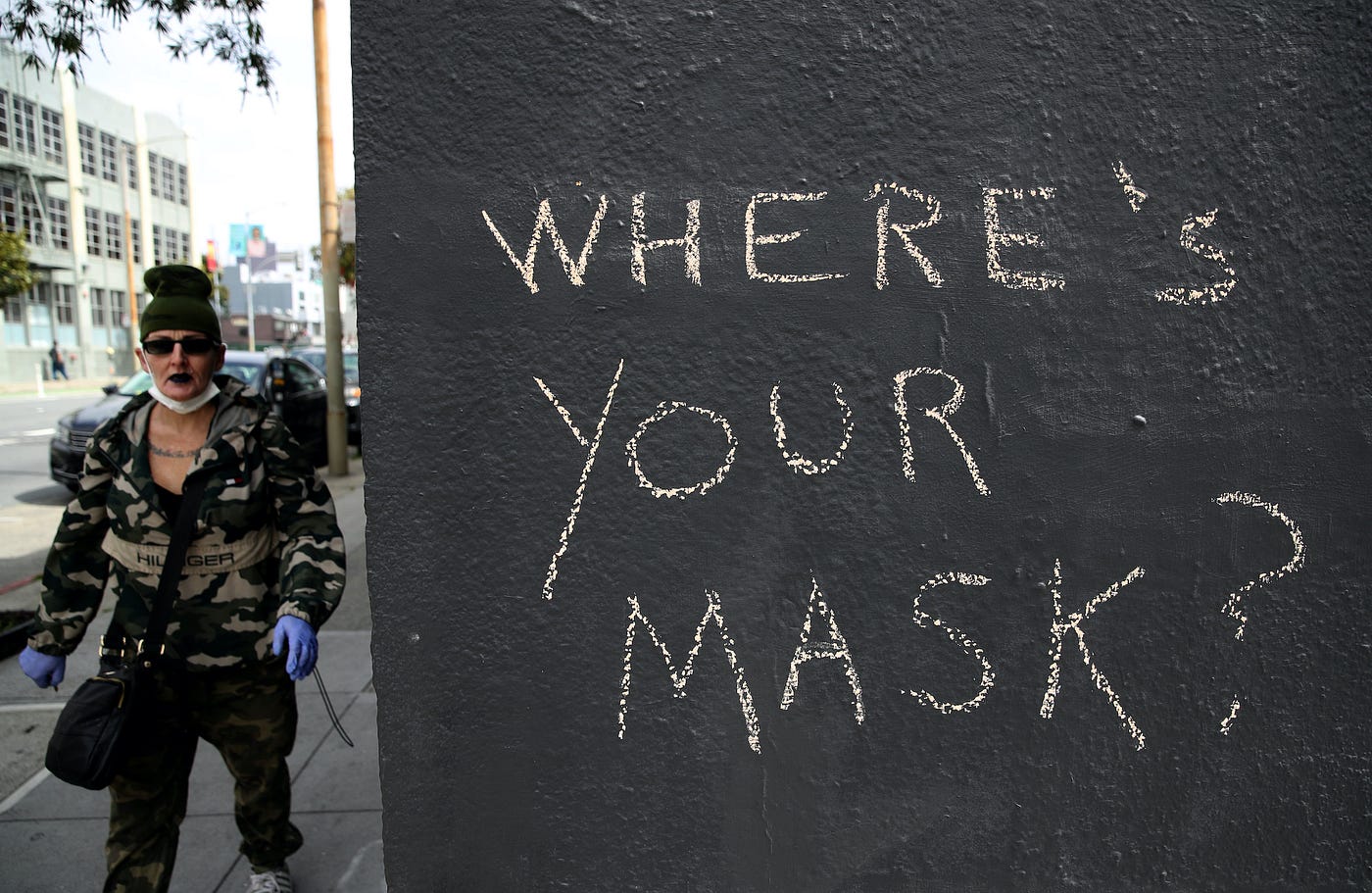 On the right side, three-quarters of the pic is a black exterior wall with “Where’s your mask?” written on it in chalk. The quarter of the photo on the left side is a person wearing latex gloves and a face mask, the latter under their chin.