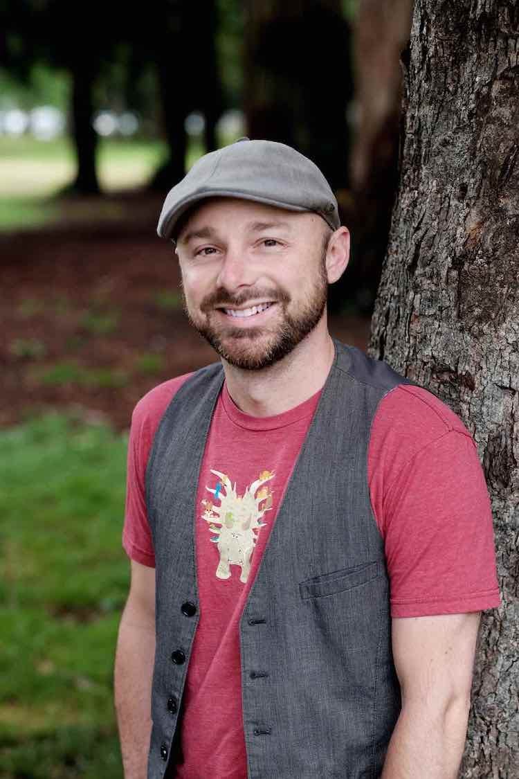 Miah, in a red t-shirt and gray vest and wearing a gray hat, smiles and poses next to a tree.