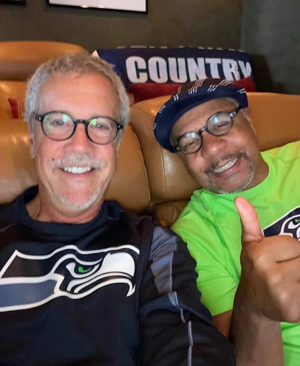A white man and a Black man, smiling and both wearing glasses and Seattle Seahawks shirts, sitting on a leather couch.