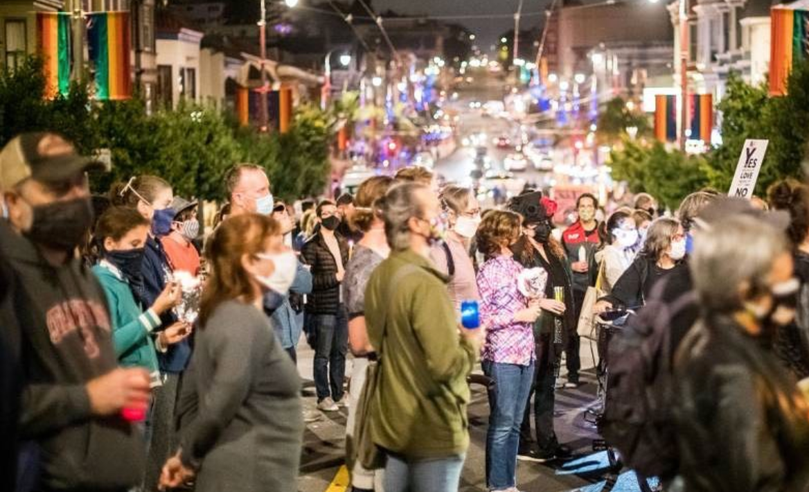 A group of people with face masks on a street, watching someone off-camera speak.