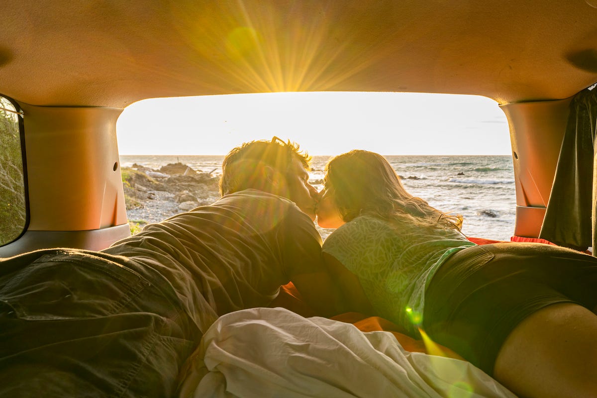 Couple lying down in a van, with the ocean in the background and the sun setting.