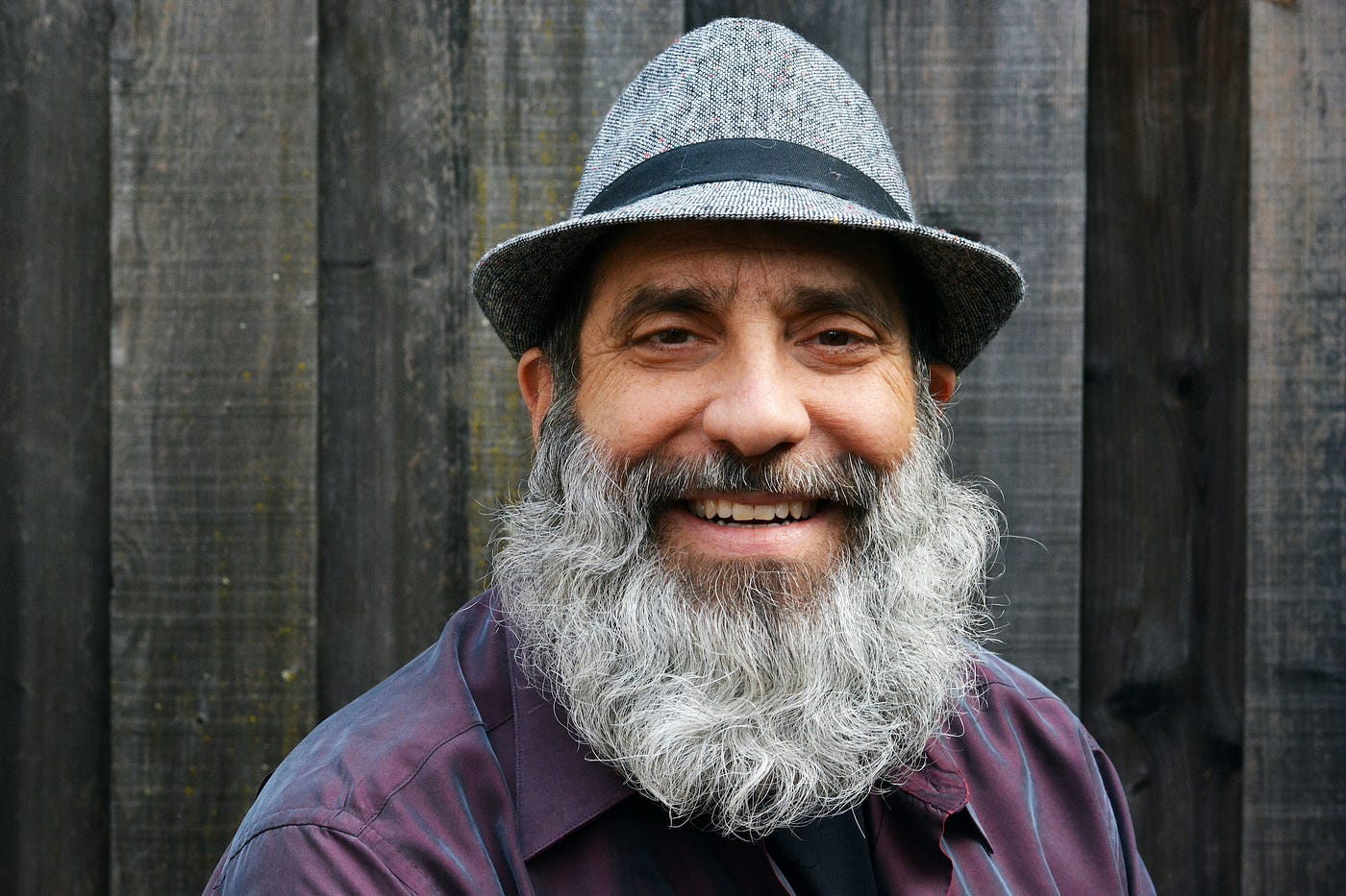 The author in front of a wooden wall. He’s wearing a hat and has a long gray beard.
