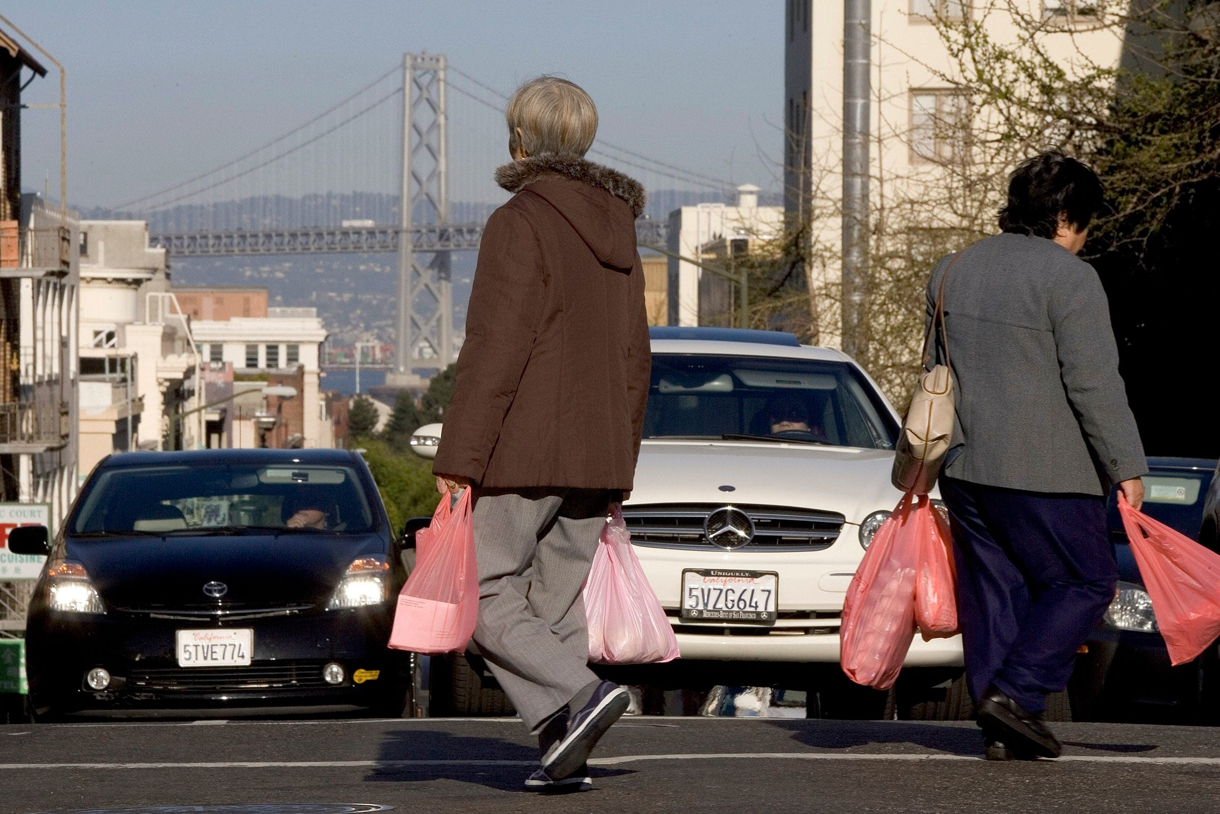 2 people carrying plastic bags crossing the street at a crosswalk at the top of a hill—the Golden Gate Bridge in the distance