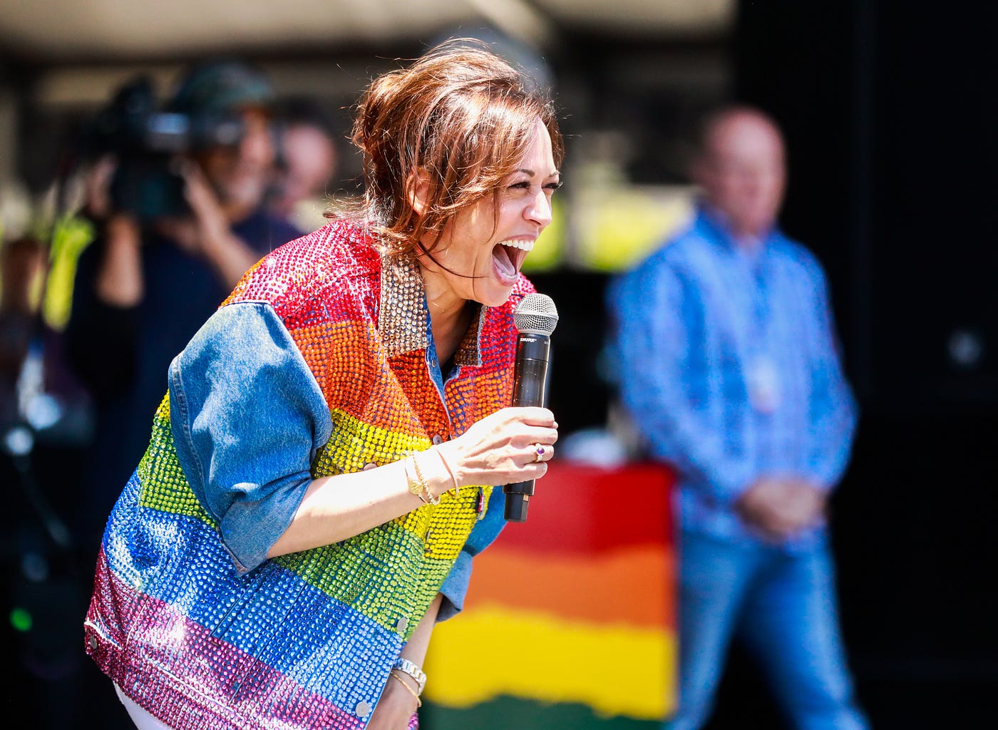 Kamala Harris speaking at an event on stage, wearing a sparkly button-up shirt with the rainbow pride flag on it