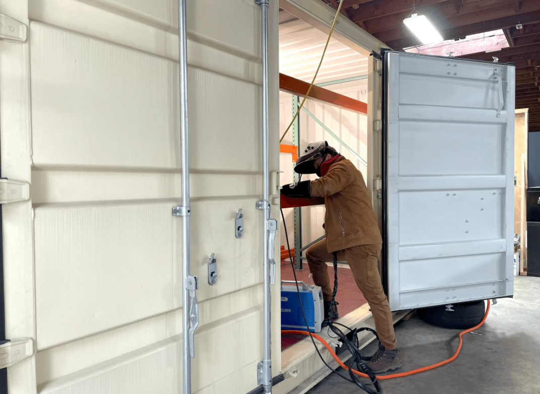 A person loading something into a metal shipping container that has only one door open, meaning we can’t see what’s inside.