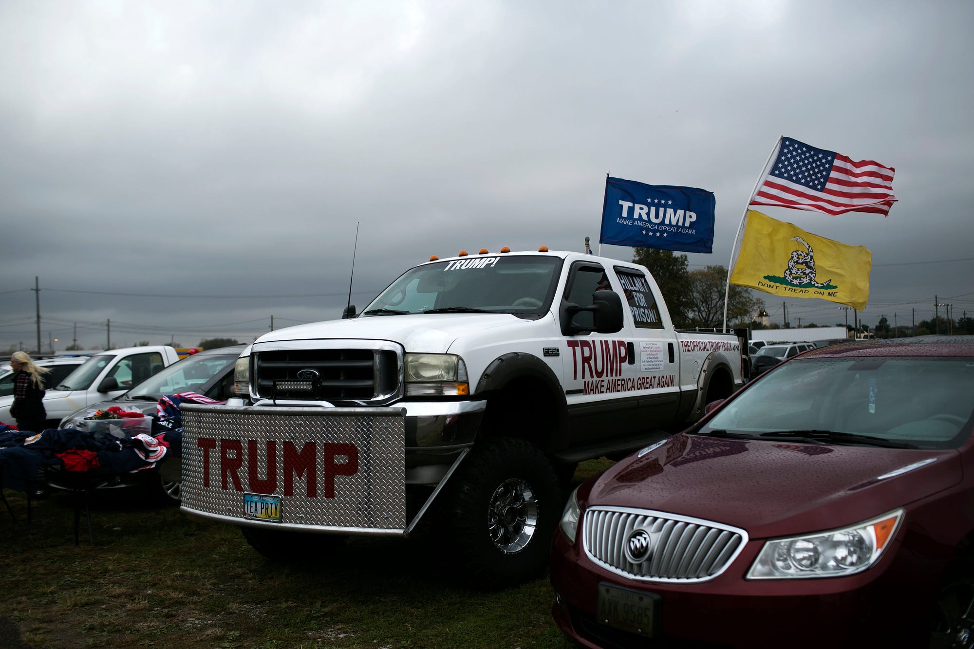A row of parked cars—a large pickup is in the center, “Trump” plastered on its doors, flying US + Trump flags.