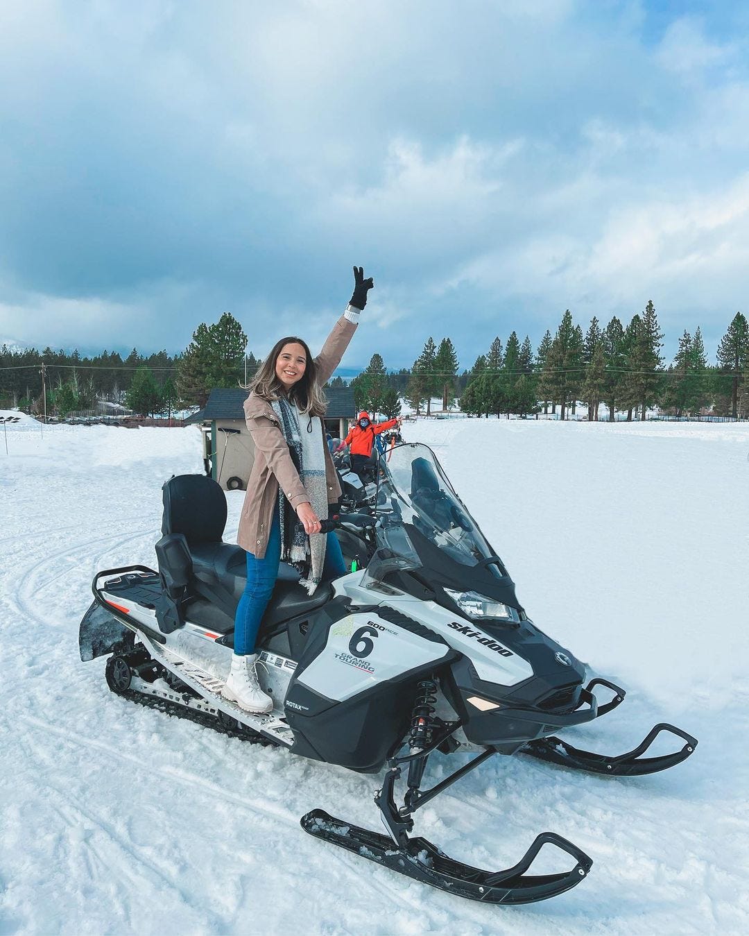 A snowmobiler gives a peace sign in the middle of a snowy field with evergreens in the background.