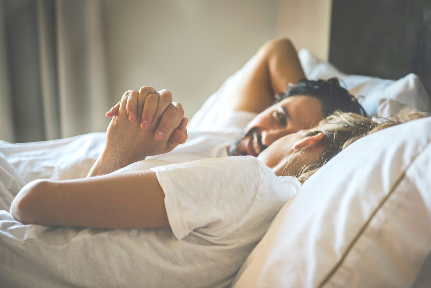 Two people lying in bed holding hands looking at each other.