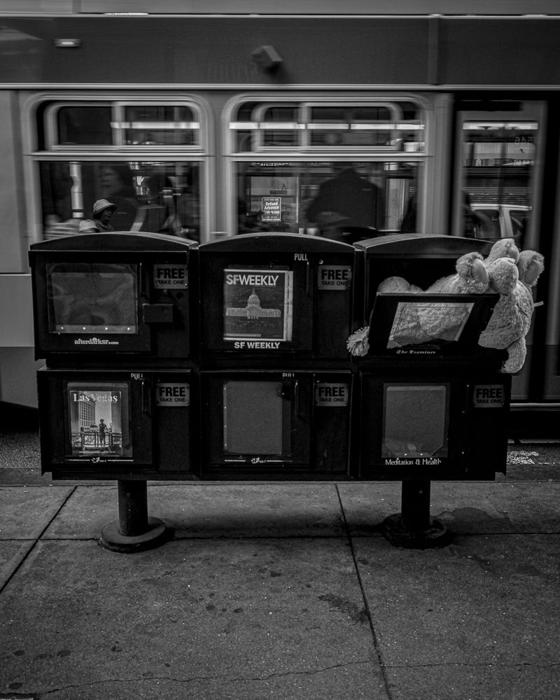 Newspaper boxes on the street, one with a stuffed animal in it, with a Muni bus in the background.