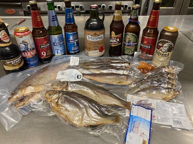 Different kinds of plastic-wrapped whole fish laid out in front of a line of imported beer in bottles and cans.