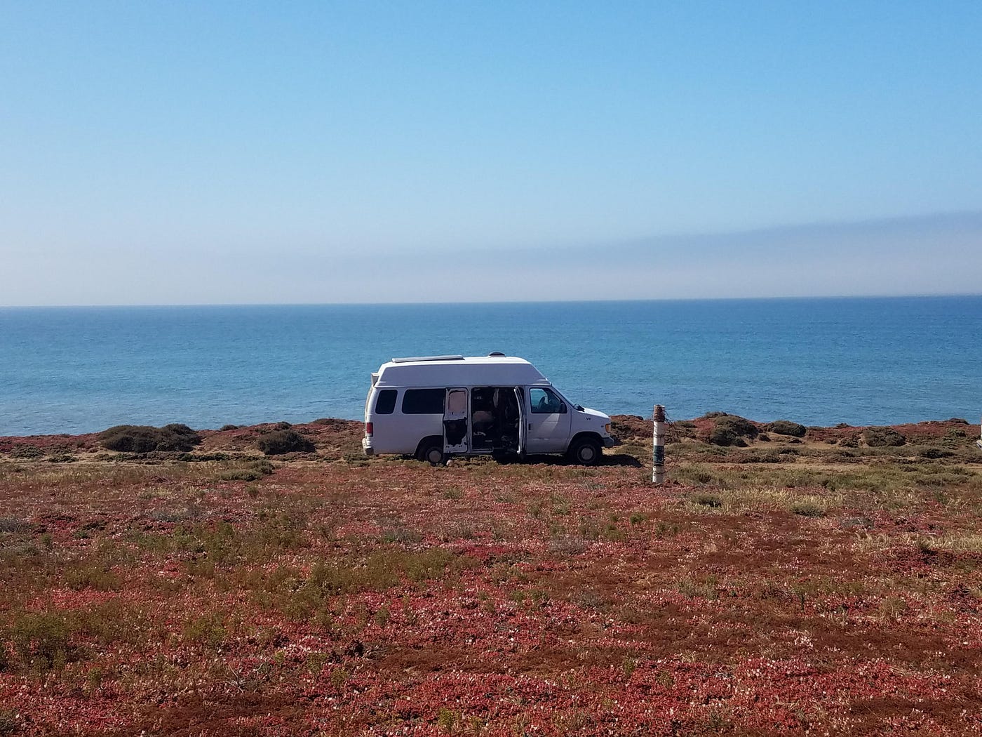 A van with its side door open, parked on grass in front of the ocean.
