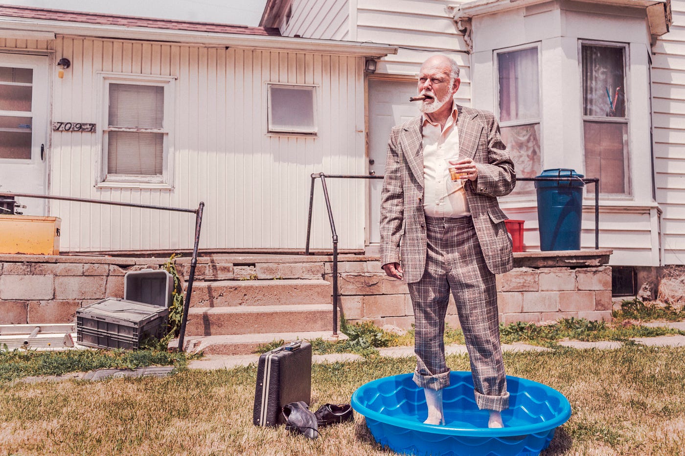 A businessman smoking a cigar, standing in a kiddie pool in front of an old house.