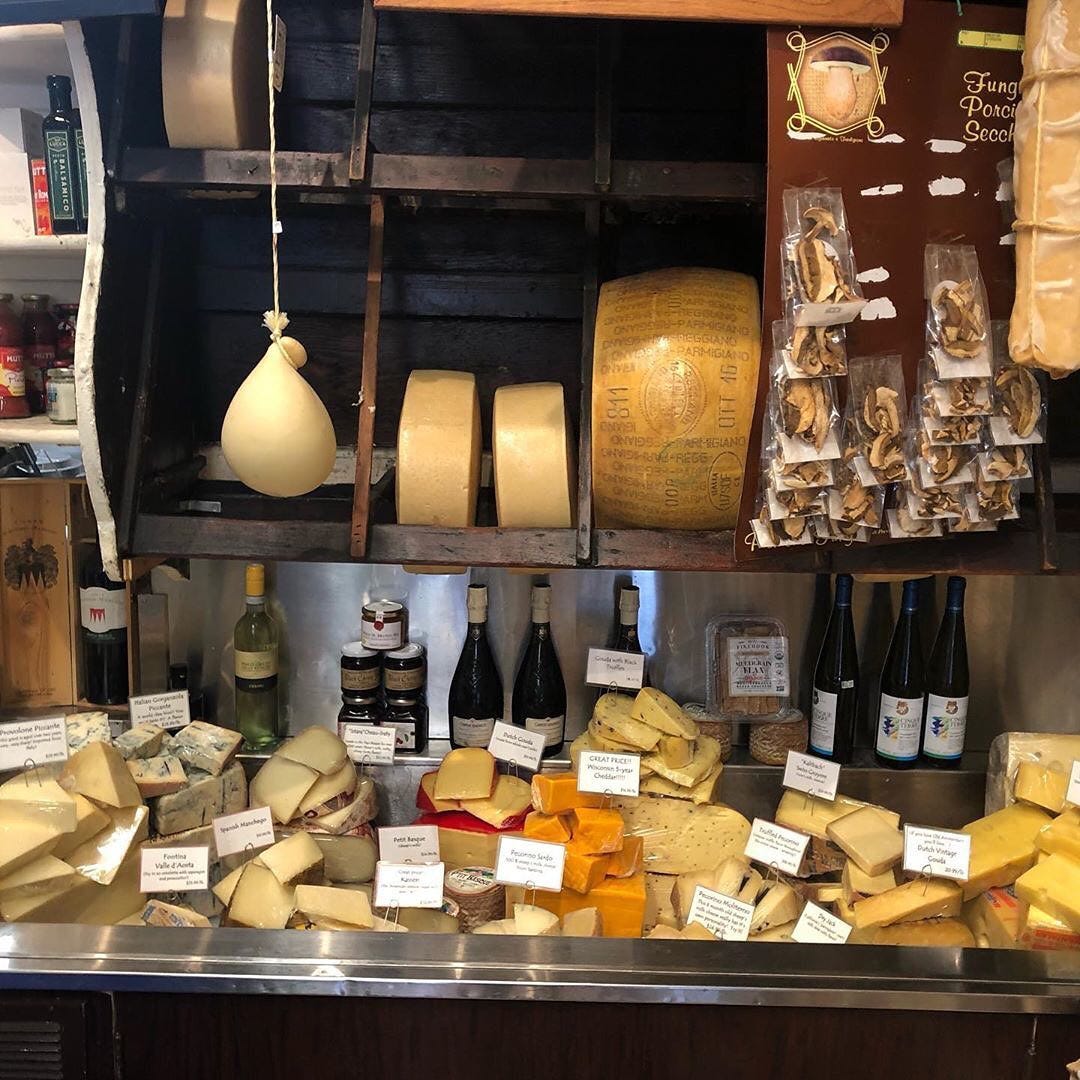 A refrigerated bin with wedges of different kinds of cheese. A shelf above it has whole wheels of cheese.