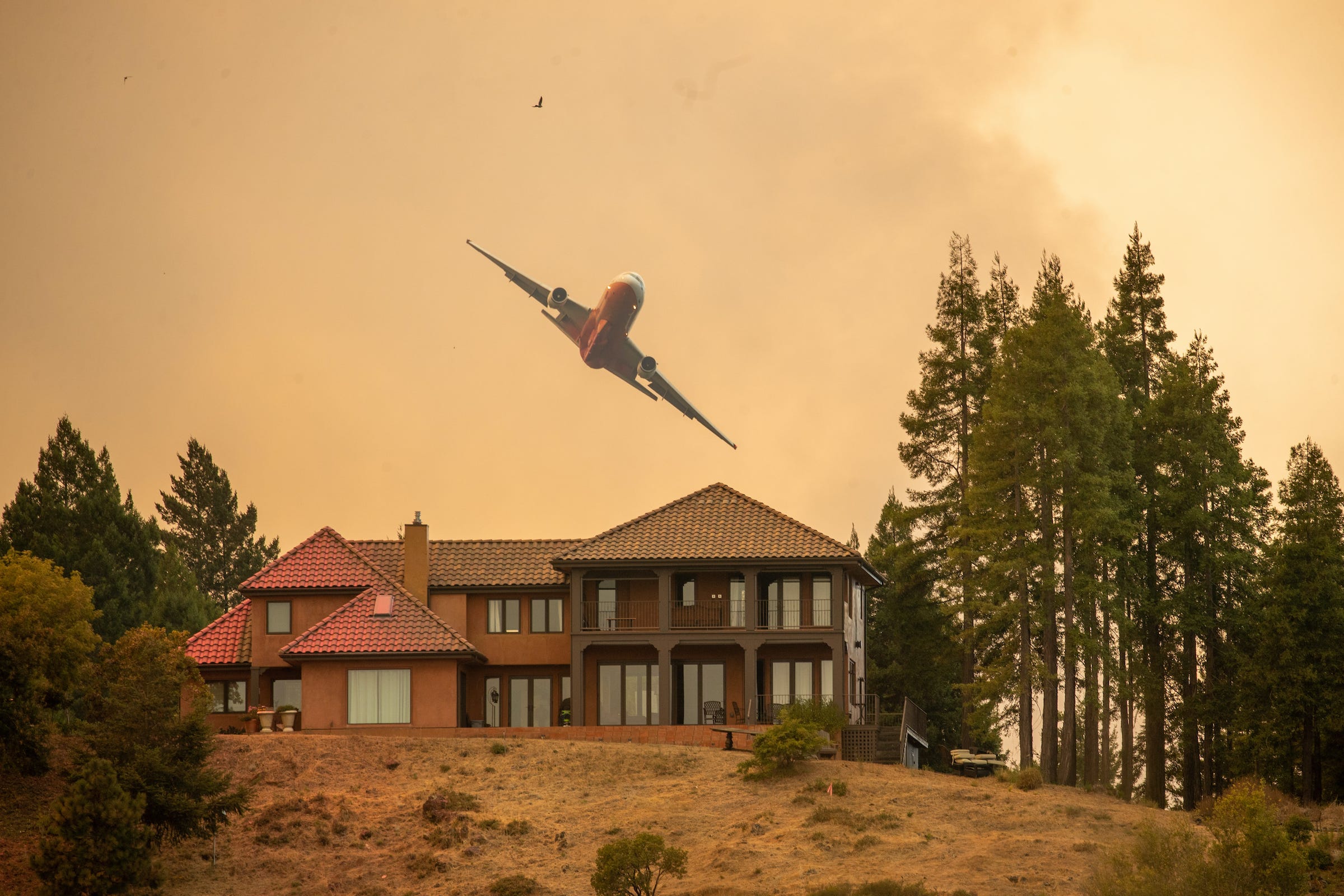 An air tanker flies in a smoky sky over a house after dropping fire retardant.