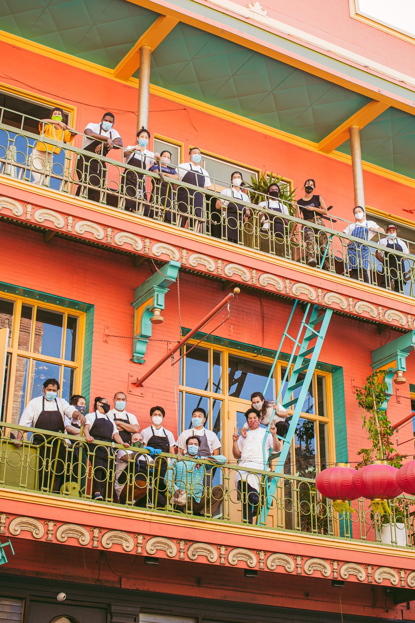 Masked staff stand on two balconies on the outside of a brightly-colored building.