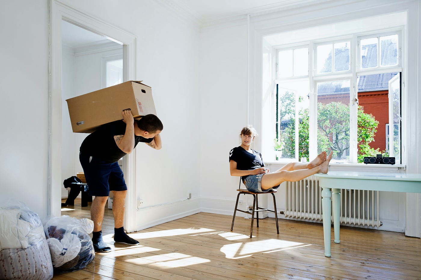 A mover carrying a box on their back, looking at a person with their feet on a table next to an open window.