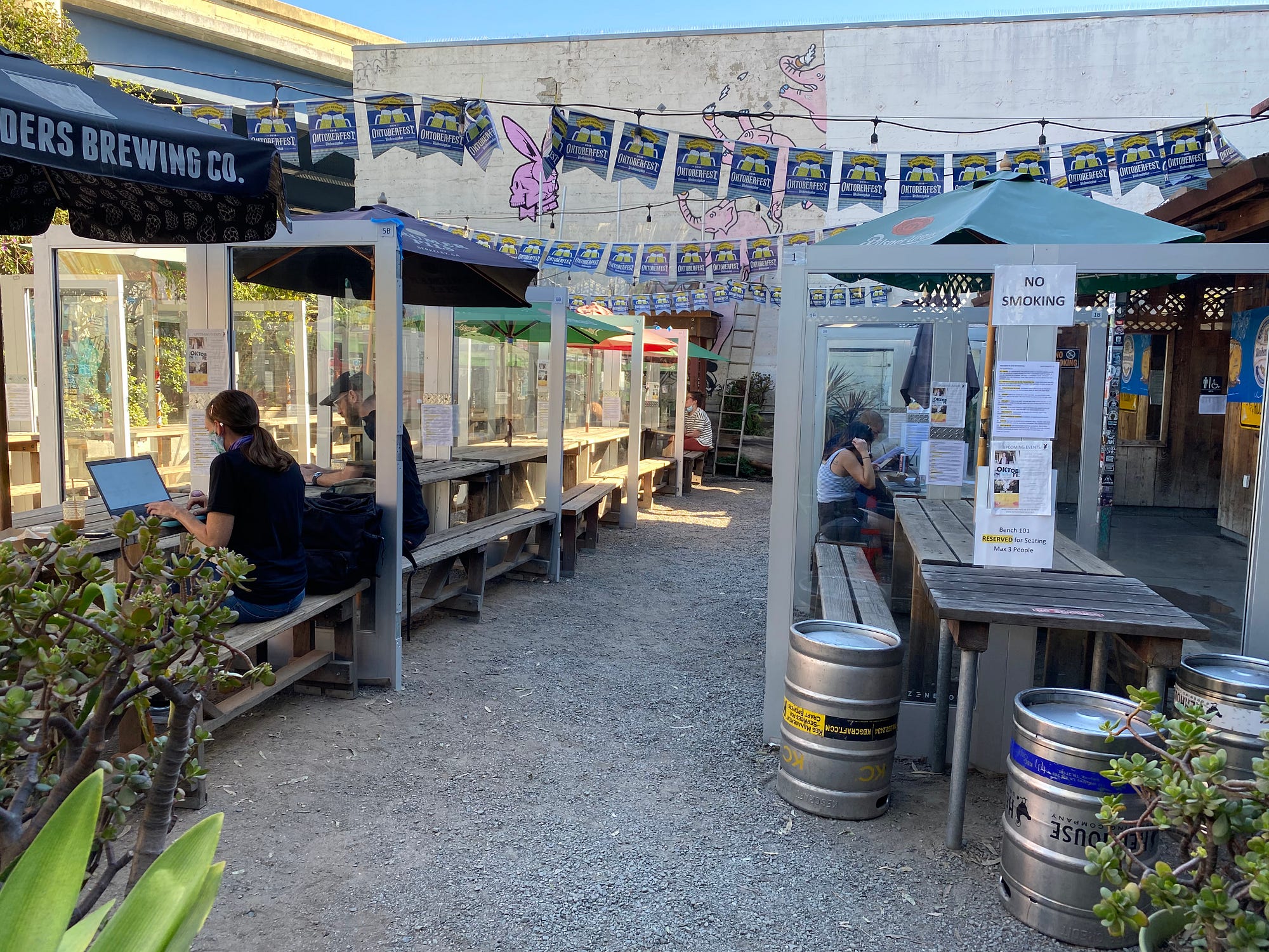 An outdoor dining area with 3 people sitting on separate wooden benches under tent covers.