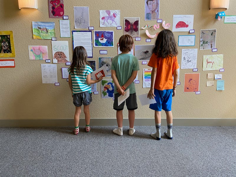 Three kids with their backs to the viewer, looking at dozens of drawings taped to the wall in front of them.