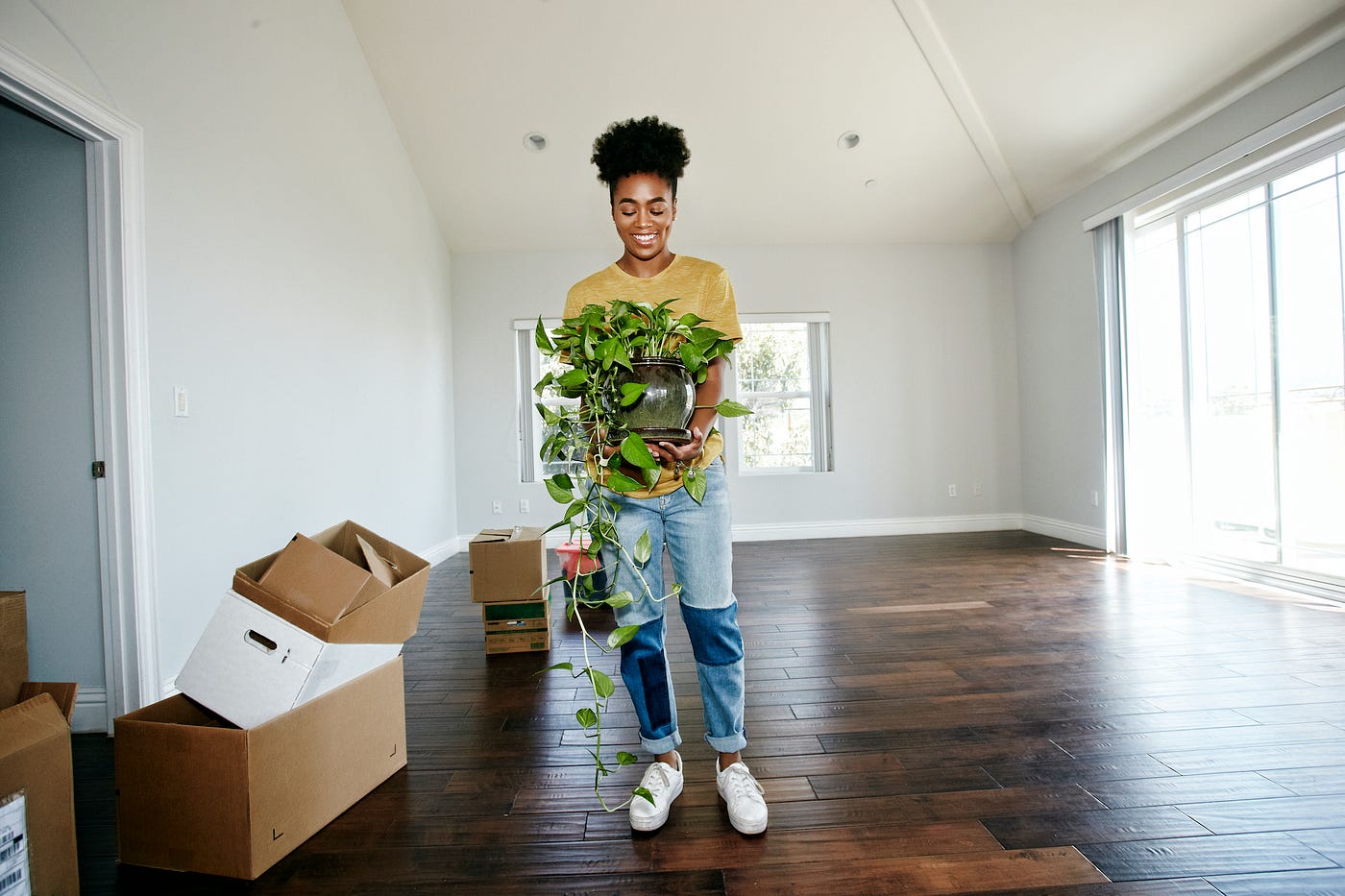 A person holding a potted plant in a living room that’s empty except for moving boxes.