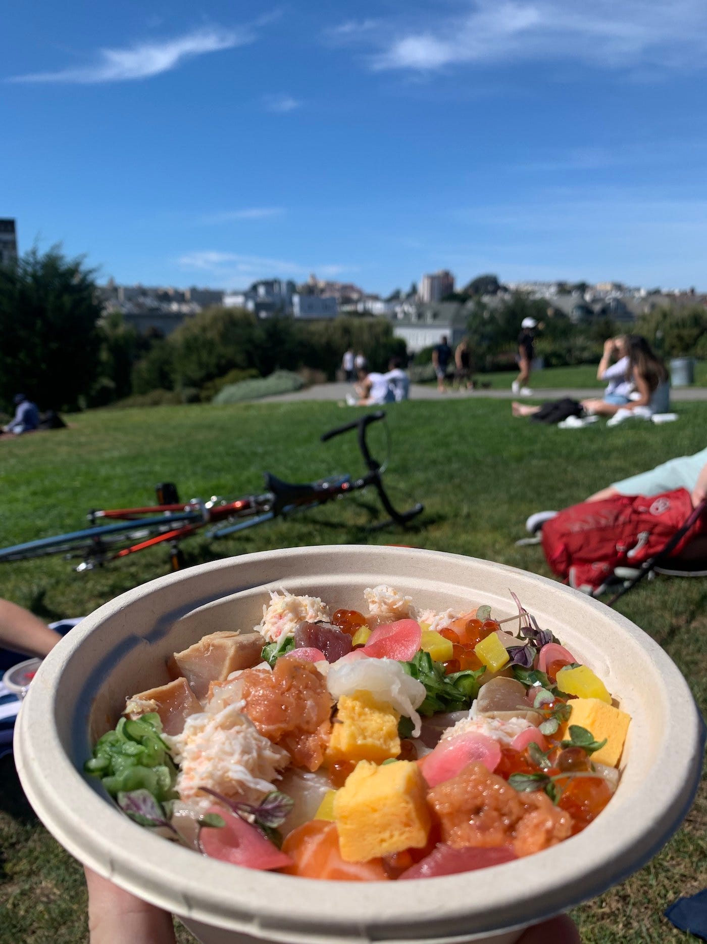 A hand holding a take-out bowl of poke with a grassy slope in the background overlooking the city. Other people are lounging around in the frame; a bicycle sits tipped on its side on the ground a few feet from the camera.