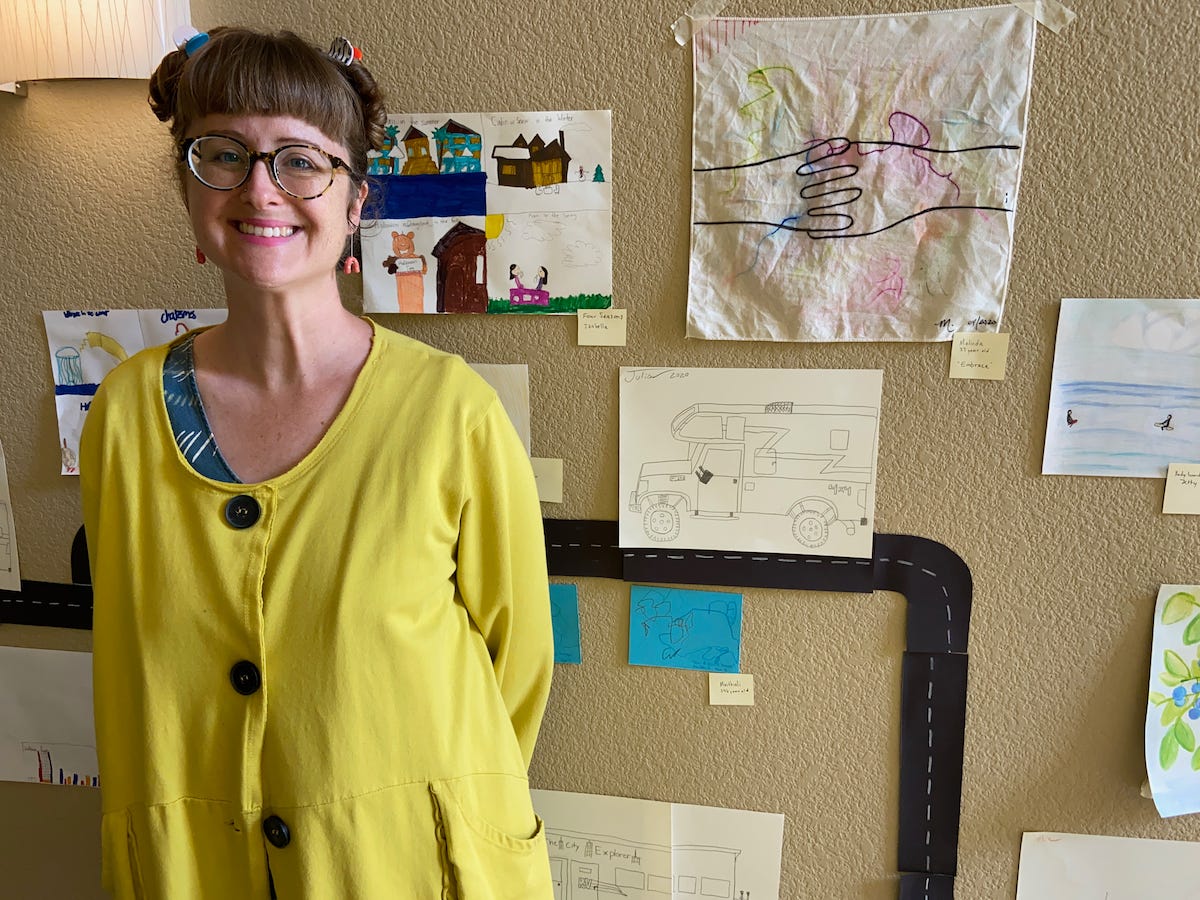 Amy Brown poses with her hands behind her back in front of her works of art posted on the wall behind her.