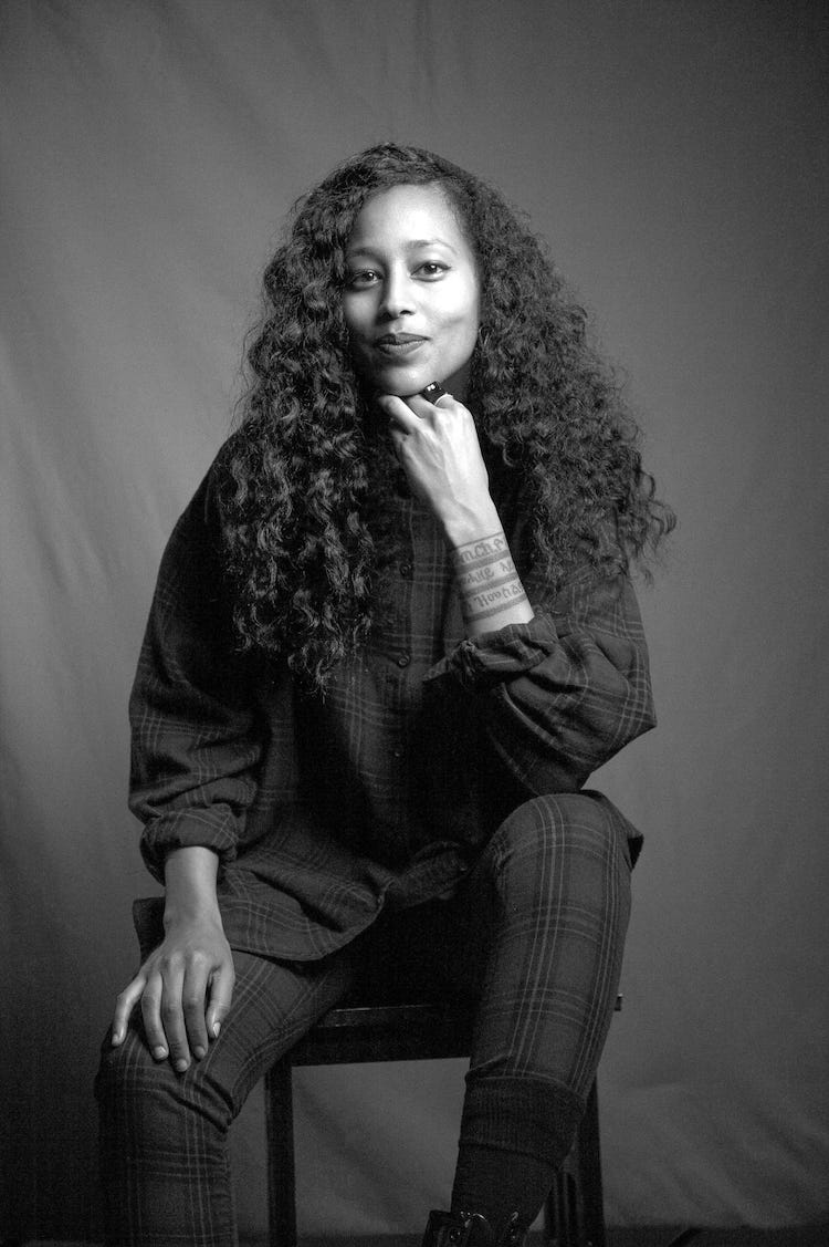 Black-and-white photo of Sephora Woldu sitting on a chair with her fist under her chin, smiling.