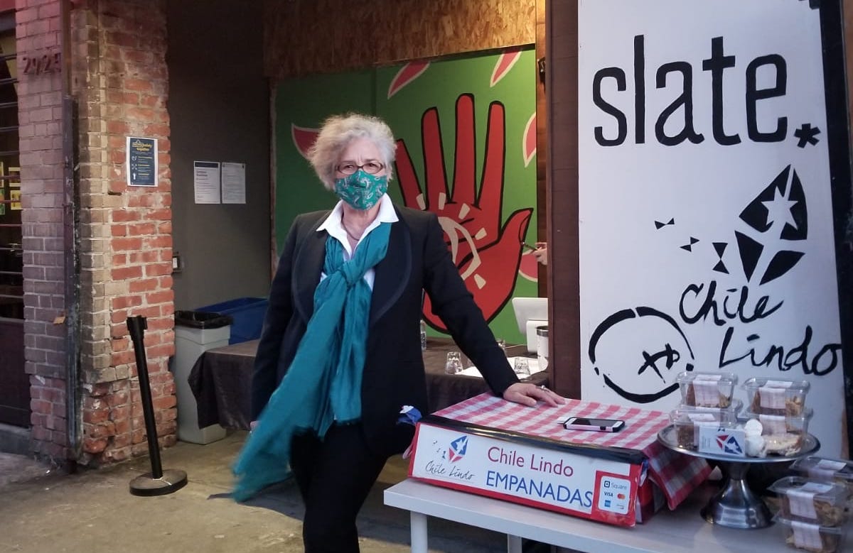 Paula Tejeda wearing a suit with a teal scarf and face mask, standing outside with a table of Chile Lindo empanadas.