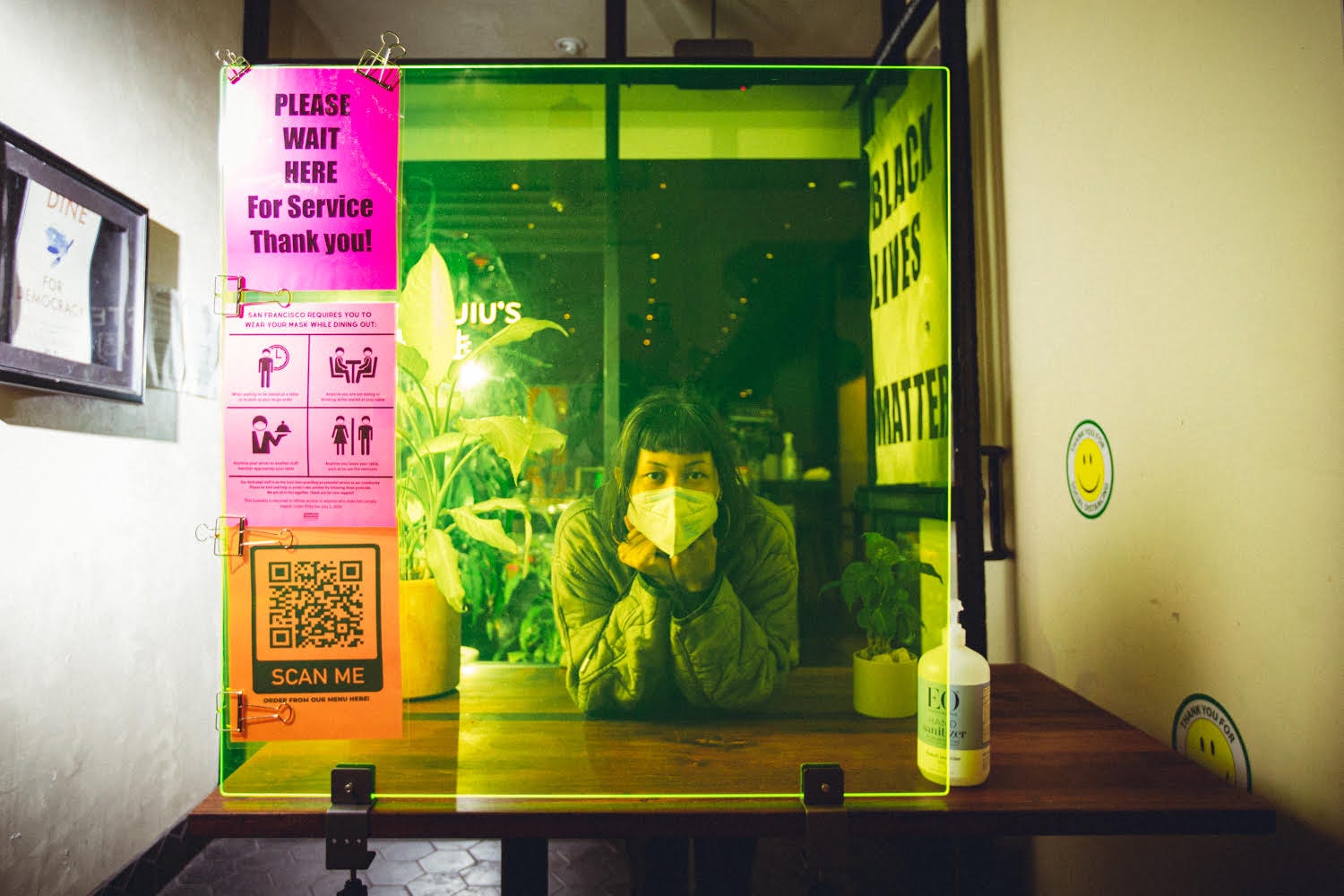 A masked hostess waiting on the other side of a neon yellow plexiglass partition at the entrance to a restaurant.