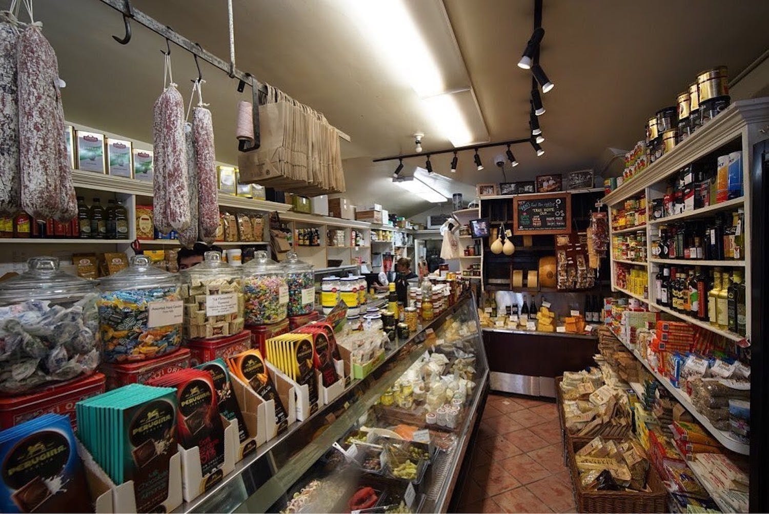 A narrow deli with a counter stocked with snacks, wine, and cheese.